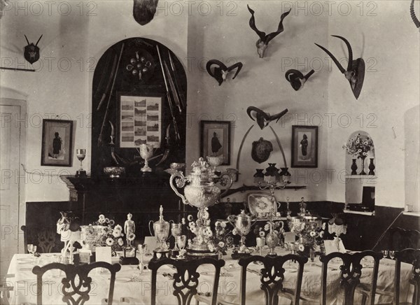 A British Army officers' dining room. Interior shot of an officers' dining room in a British Army mess building. Framed pictures and mounted antelope skulls adorn the walls around a table set with fine china, flowers and silverware. Several silver trophies make up a centrepiece. Chaman, Baluchistan, India (Balochistan, Pakistan), 1905. Chaman, Balochistan, Pakistan, Southern Asia, Asia.