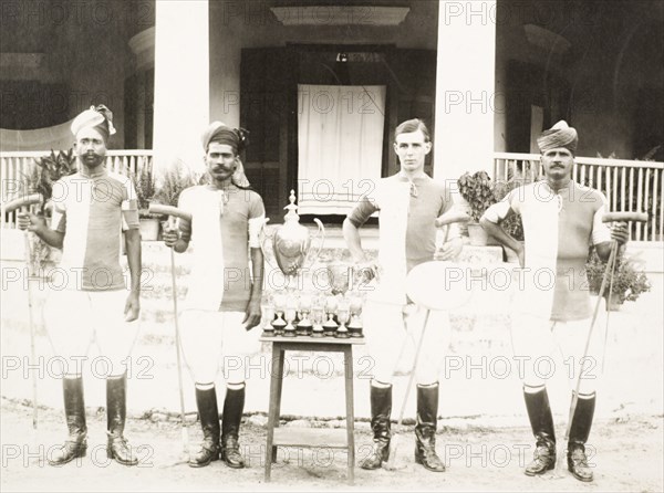 An Indian Police polo team. Members of an Indian Police polo team pose for a group portrait beside a table covered with trophies. The four men (three Indian and one British) stand, holding their polo sticks, dressed in matching polo shirts, breeches and riding boots. India, circa 1927. India, Southern Asia, Asia.