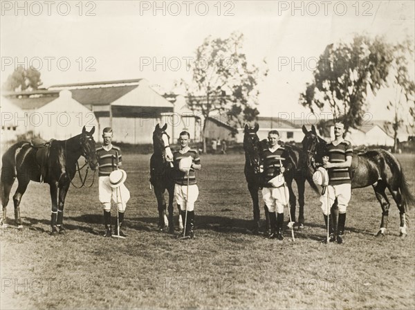 Polo players with their horses, Kenya. Four polo players, probably members of the Kenya Police team, pose outdoors with their horses and polo sticks. They stand, holding their hats, dressed in matching striped polo shirts, breeches and riding boots. Kenya, circa 1927. Kenya, Eastern Africa, Africa.