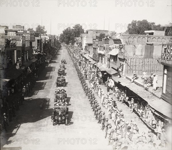 Indian Army officers salute a procession of cars. Hundreds of Indian Army officers, and crowds of civilians, line a city street as a procession of chauffeur-driven cars passes by. Each vehicle contains several British men in uniform, probably government or military officials on their way to a formal engagement. Probably northern India, circa 1923. India, Southern Asia, Asia.