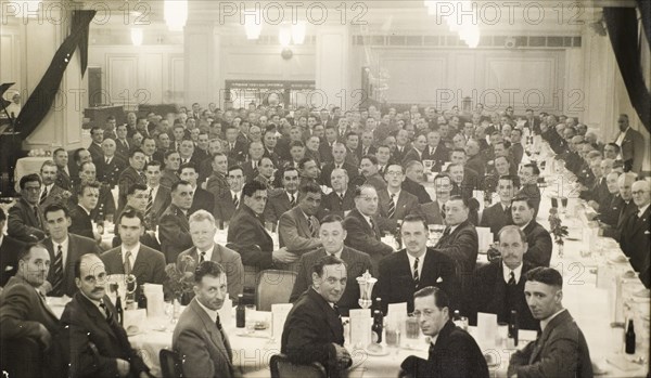 Colonial Police Force dinner, 1954. A large group of British men attend a formal dinner and award ceremony for members of the Colonial Police Force. They sit around long banquet tables in a dining hall, dressed in suits and matching ties. Two members of the Nigeria Police Force sit in the front row: Ian S. Proud, Assistant Commissioner in Lagos (third from left), and Kerr Bovell, Inspector General (fourth from left). Probably London, United Kingdom, 1954. London, London, City of, England (United Kingdom), Western Europe, Europe .