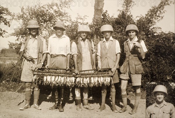 Trophies from a snipe hunt. Five British hunters stand side-by-side, displaying the trophies of a recent snipe hunt for the camera. The dead birds hang by their necks from wooden racks. A portrait of another man appears in the corner, perhaps a member of the hunting party missing from the photograph. Probably Barisal Division, India (Bangladesh), 31 October 1937. Bangladesh, Southern Asia, Asia.