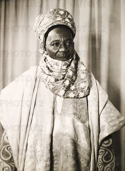 Portrait of a Nigerian man in traditional dress. Portrait of a Nigerian man dressed in fine clothes, probably a chief or dignitary. He wears a 'buba' (loose fitting shirt) beneath a patterned 'agbada' (overgarment), with a headscarf wrapped around his head and neck. Nigeria, circa 1955. Nigeria, Western Africa, Africa.