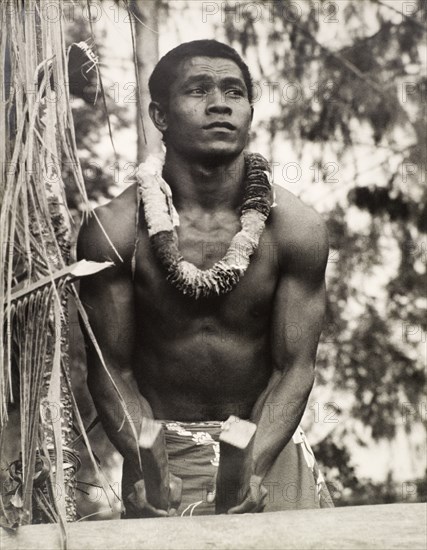 A secondary school student, Solomon Islands. A student of the King George VI School in Auki, drums using two roughly chopped pieces of wood as drumsticks. He is naked from the waist up and wears a floral garland around his neck. Auki, Malaita Island, Solomon Islands, circa 1960. Auki, Malaita, Solomon Islands, Pacific Ocean, Oceania.