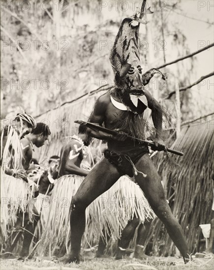 Student performing in traditional costume, Solomon Islands. A student of the King George VI School in Auki, wears traditional costume for a performance. He is dressed in an elaborate, painted mask, which covers his head, and holds a short spear with both hands. Auki, Malaita Island, Solomon Islands, circa 1960. Auki, Malaita, Solomon Islands, Pacific Ocean, Oceania.