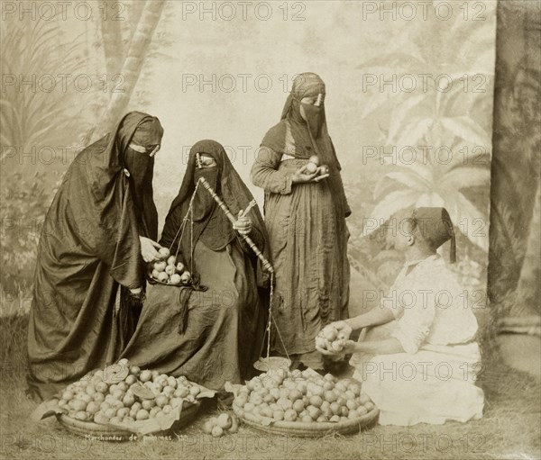 North African apple seller. Posed studio portrait of a man wearing a fez selling apples to three heavily veiled Muslim women. The seller sits on the ground beside his baskets whilst two of the women place apples in a balance. The group are arranged against a backdrop cloth painted with exotic foliage and the ground appears to have been spread or planted with grass, a common feature in studio photographs of racial types. Probably Egypt, circa 1895. Egypt, Northern Africa, Africa.