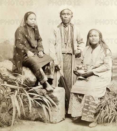 A 'Malay Hadjee' and family. Studio portrait of a 'Malay Hadjee' and family. The man wears traditional dress with a white turban indicating that he has completed the Hajj or Islamic pilgrimage to Mecca. He has a pocket watch and a Western umbrella. Two women, representing his wife and daughter, wear traditional Malay dress or 'baju kurung', a loose tunic worn over a sarung or sarong. Each wears an embroidered 'tudung' or headscarf, the older woman holding a set of utensils for preparing 'siri' or betel nut. Singapore, Straits Settlements (Singapore), circa 1900. Singapore, South East Asia, Asia.