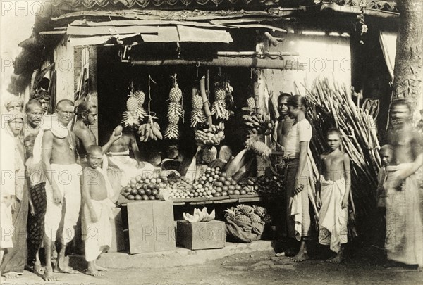 Ceylonian fruit stall. A group of Ceylonian men, women and children in traditional dress stand around a fruit-seller's stall that is festooned with pineapples. Ceylon (Sri Lanka), circa 1900. Sri Lanka, Southern Asia, Asia.
