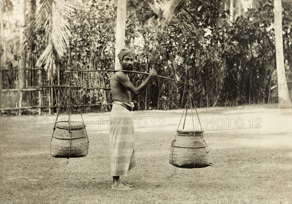 Ceylonian man with baskets. A Ceylonian man, possibly a snake charmer, carries two baskets on a pole across his shoulder. Ceylon (Sri Lanka), circa 1900. Sri Lanka, Southern Asia, Asia.