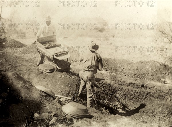 Digging for gold at Kalgoorlie. Two miners dig for gold in the outback: one man shovels earth from a shallow pit onto a gold shaker operated by the other. The machine was designed to sift through dirt and gravel, carrying the heavier gold to the bottom. Probably Kalgoorlie, Australia, circa 1901. Kalgoorlie, West Australia, Australia, Australia, Oceania.