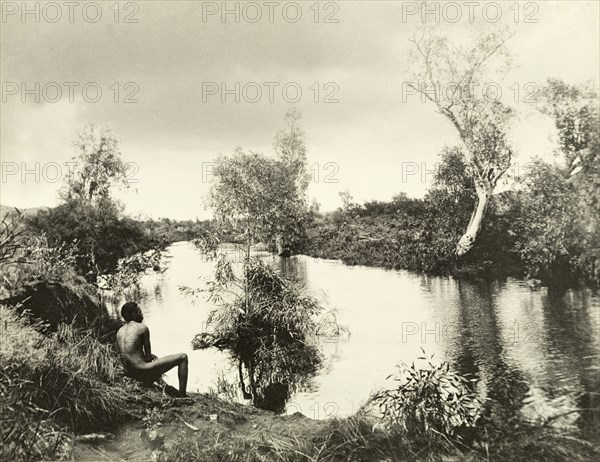An aborigine on the banks of a backwater. An aborigine sits with his back to the camera on the banks of a peaceful backwater in the outback. Western Australia, Australia, circa 1901., West Australia, Australia, Australia, Oceania.