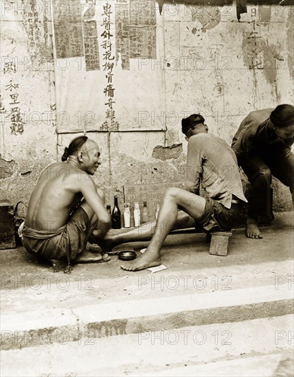 A medicine stall in Hong Kong. A man wearing a traditional Manchu hairstyle squats beside a row of glass bottles, chatting to a medicine man at a street stall in Hong Kong. Hong Kong, China, circa 1903. Hong Kong, Hong Kong, China, People's Republic of, Eastern Asia, Asia.