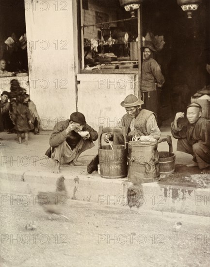 A meal by the wayside', Hong Kong. A Chinese street trader distributes food from a wooden bucket by the roadside. A customer squats down beside him, eating from a bowl with chopsticks. Hong Kong, China, circa 1903. Hong Kong, Hong Kong, China, People's Republic of, Eastern Asia, Asia.