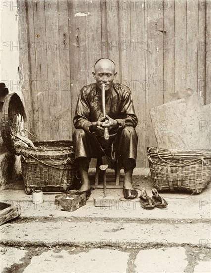 An itinerant shoemaker, Hong Kong. An itinerant shoemaker sits on a stool, smoking a long pipe at his street stall in Hong Kong. A shoe mould, a box of tools and a pair of sandals are positioned in front of him. Hong Kong, China, circa 1903. Hong Kong, Hong Kong, China, People's Republic of, Eastern Asia, Asia.
