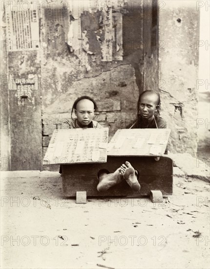 Petty criminals in the stocks, Hong Kong. Two petty criminals sit with their ankles restrained in stocks, suffering public humiliation on a city street. Each wears a board strung around his neck, describing in detail the offense committed. Hong Kong, China, circa 1903. Hong Kong, Hong Kong, China, People's Republic of, Eastern Asia, Asia.