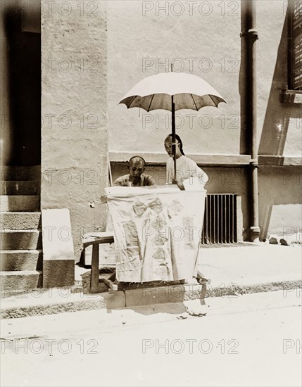 Letter-writing service on a Hong Kong street. A street trader offering a letter-writing service sits beneath a parasol at his makeshift stall. Hong Kong, China, circa 1903. Hong Kong, Hong Kong, China, People's Republic of, Eastern Asia, Asia.