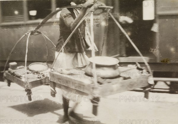 Burmese hawker at a railway station. A Burmese hawker attempts to sell his wares to passers-by at a railway station. He carries various pots, probably containing food, on two large trays suspended from a pole across his shoulder. Probably Mandalay, Burma (Myanmar), circa April 1921., Mandalay, Burma (Myanmar), South East Asia, Asia.