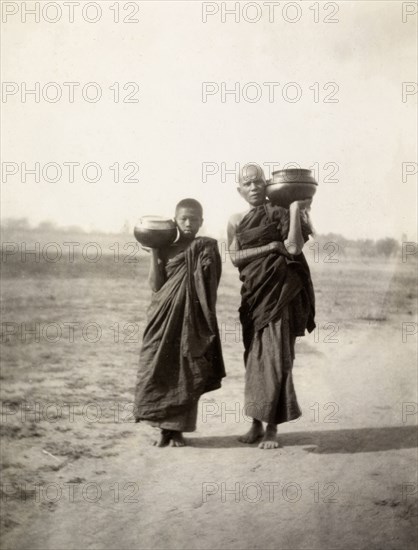 Pot carriers in Mandalay. A Burmese man and a boy dressed in long robes carry pots on their shoulders along a dusty road at Fort Dufferin. Mandalay, Burma (Myanmar), circa February 1927. Mandalay, Mandalay, Burma (Myanmar), South East Asia, Asia.