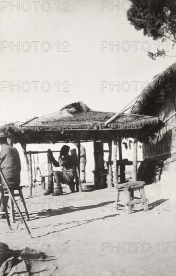 On watch at 'Pagoda Hill'. A Burmese man dressed in military uniform and a fez hat sits on a stool beneath a thatched canopy, peering through a long telescope. An original caption gives the location as 'Pagoda Hill', possibly a reference to the Kakku Pagodas located near Taunggyi. Taunggyi, Burma (Myanmar), December 1928. Taunggyi, Shan, Burma (Myanmar), South East Asia, Asia.