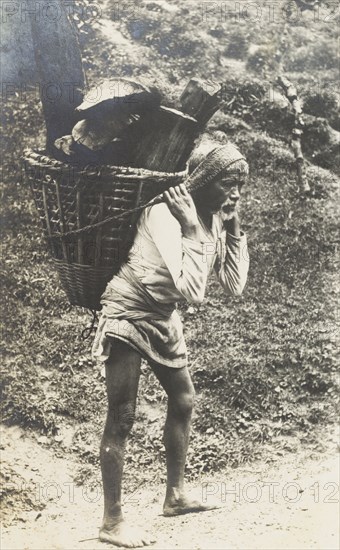 Transporting firewood in the hills. Portrait of an elderly man carrying a basket full of firewood in the hills. Hunched over and barefoot, he bears the load with the aid of a strap attached to his head. Probably Nepal, circa 1922. Nepal, Southern Asia, Asia.