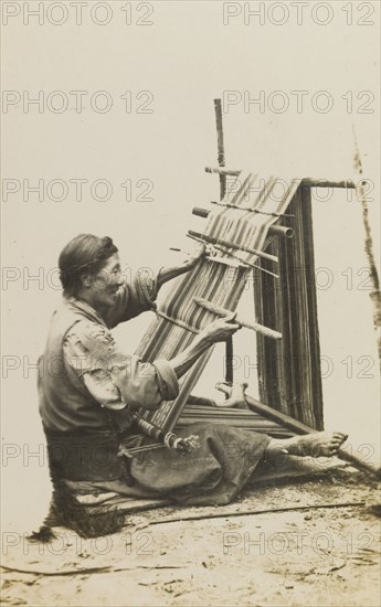 Bhatia woman weaving cloth. An elderly Bhatia woman sits on the ground, operating a frame loom by hand to weave a length of patterned cloth. India, circa 1922. India, Southern Asia, Asia.