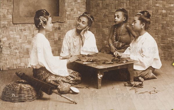 Burmese women rolling cheroots. Three young Burmese women kneel around a low table as they roll 'cheroots' (cigars). Dressed in loose-fitting shirts and traditional 'longyis', they wear their hair tied up and decorated with flowers. They are accompanied by a young child who watches with interest. Burma (Myanmar), circa 1926. Burma (Myanmar), South East Asia, Asia.