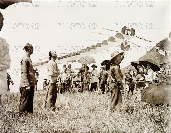 Flying a dragon kite. A long kite, constructed in segments to look like a dragon, is made ready to fly. A group of spectators gather to watch, sheltering from the sun beneath parasols. China, circa 1905. China, People's Republic of, Eastern Asia, Asia.