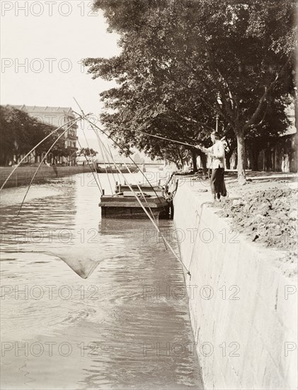 Fishing in Bowrington Canal. A Chinese fisherman raises his net from Bowrington Canal, pulling on a rope attached to a light, wooden frame to lift it from the water. Bowrington Bridge is visible in the distance. Wan Chai, Hong Kong, China, circa 1905. Wan Chai, Hong Kong, China, People's Republic of, Eastern Asia, Asia.