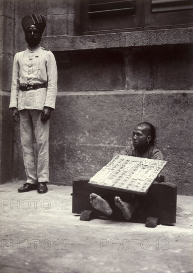 Petty criminal in the stocks, Hong Kong. A petty criminal sits with his ankles restrained in stocks, guarded by an Indian officer from the Hong Kong Police Force. He wears a board strung around his neck, describing in detail the offense committed. Hong Kong, China, circa 1905. Hong Kong, Hong Kong, China, People's Republic of, Eastern Asia, Asia.
