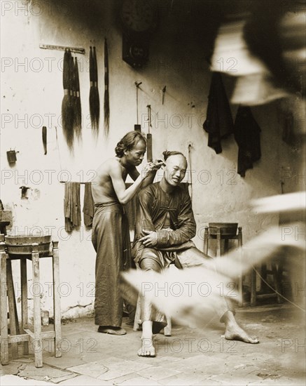Ear cleaning at a barber's shop, China. A man with a distinctive Manchu hairstyle sits on a stool to have his ears cleaned at a barber's shop. Several false 'queues' (waist-long pigtails), hang on the wall behind him: an equivalent to modern-day hair extensions. China, circa 1905. China, People's Republic of, Eastern Asia, Asia.