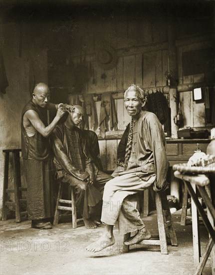 Ear cleaning at a barber's shop, China. A man sits on a stool to have his ears cleaned at a barber's shop. An elderly man with a long, plaited 'queue' (waist-long pigtail) sits nearby, waiting his turn. China, circa 1905. China, People's Republic of, Eastern Asia, Asia.