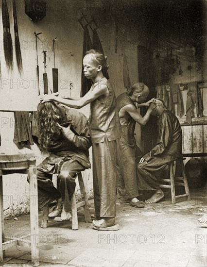 Inside a Chinese barber's shop. Two men with distinctive Manchu hairstyles receive treatment at a barber's shop. One sits with his long hair thrown forward whilst a barber shaves the back of his head: the other tilts his head to have his ears cleaned. Several false 'queues' (waist-long pigtails), hang on the wall behind them: an equivalent to modern-day hair extensions. China, circa 1905. China, People's Republic of, Eastern Asia, Asia.