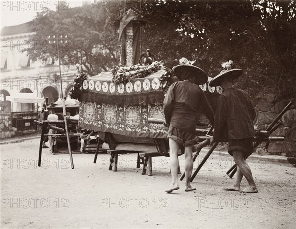 Pallbearers at a Chinese funeral. Two pallbearers prepare to carry a coffin on a wooden bier during a Chinese funeral ceremony. The coffin is draped in fine cloth and decorated with wreaths of flowers, indicating that the deceased had been wealthy. China, circa 1905. China, People's Republic of, Eastern Asia, Asia.