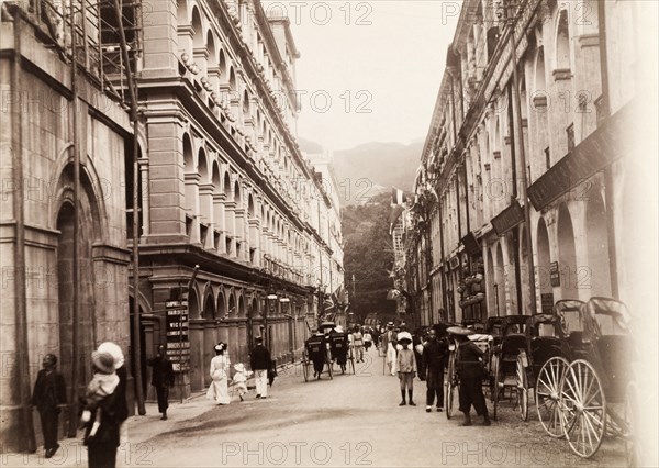 Queen's Road Central, Hong Kong. View along Queen's Road Central, looking east. Multi-storey colonial buildings line the street, which is dotted with pedestrians and rickshaws. An original caption points out the base of the Pedder Street clock tower on the left, a structure that was demolished in 1908. China, April 1910. Hong Kong, Hong Kong, China, People's Republic of, Eastern Asia, Asia.