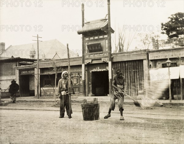 Watering the roadway in Hong Kong. A man throws water from a bucket onto the road, probably to control surface dust. Behind him, to the left, is the London Mission building. Hong Kong, China, circa 1910. Hong Kong, Hong Kong, China, People's Republic of, Eastern Asia, Asia.