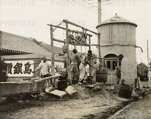 A hand-drawn street well in China. Two men collect water from a street well, carrying it away in a large wooden bucket suspended from a pole. Another man raises water from the well using a manual pulley system attached to a wooden frame. Probably Hong Kong, China, circa 1910. China, People's Republic of, Eastern Asia, Asia.