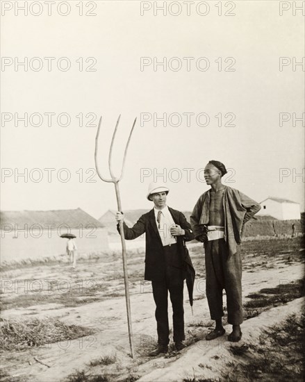 European man with Chinese farming tool. A European man, dressed in a suit and solatopi hat, holds a large, three-pronged fork as he chats to a Chinese farm labourer. Northern China, circa 1920. China, People's Republic of, Eastern Asia, Asia.