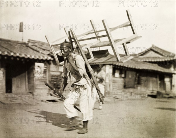 A Korean labourer carries a bench. A Korean labourer carries a wooden bench strapped to his back along a city street. Seoul, Korea (South Korea), circa 1920. South Korea, Eastern Asia, Asia.
