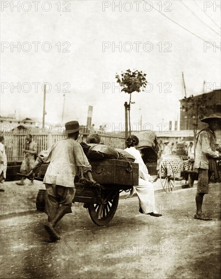 Handcart on a harbourside road in Seoul. A man pushes a handcart laden with luggage along a harbourside road in Seoul. Two women in traditional Korean dress hitch a ride on the front. Seoul, Korea (South Korea), circa 1920. Seoul, Special City, South Korea, Eastern Asia, Asia.