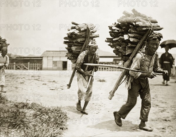 Korean labourers transporting firewood. Two Korean labourers carry large bundles of firewood, strapped to their backs on triangular wooden frames. Seoul, Korea (South Korea), circa 1920. South Korea, Eastern Asia, Asia.