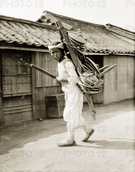 A Korean peddler boy. A Korean peddler boy carries his wares, probably crockery of some kind, on a wooden frame strapped to his back. Seoul, Korea (South Korea), circa 1920. Seoul, Special City, South Korea, Eastern Asia, Asia.