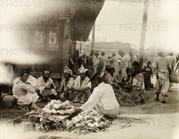 Vegetable stall at an outdoor market in Seoul. A street trader wearing a 'gat' (a traditional Korean hat), crouches down to arrange the vegetables on his stall at a busy outdoor market. Other traders and customers mill about in the background. Seoul, Korea (South Korea), circa 1920. Seoul, Special City, South Korea, Eastern Asia, Asia.
