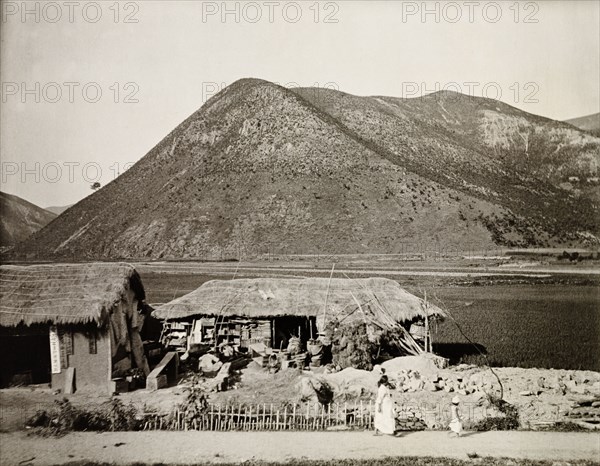 Dwelling in the mountains, Korea. A small dwelling with a thatched roof sits at the edge of a flat plain against a mountainous backdrop. Korea (South Korea), circa 1920. South Korea, Eastern Asia, Asia.