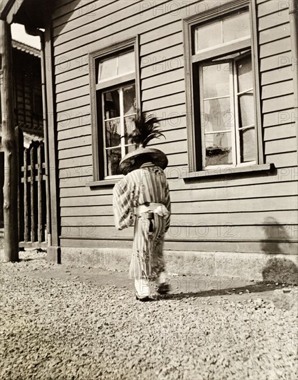 Traditional Japanese dress. A person (probably male) dressed in traditional Japanese costume, walks away from the camera. He wears a striped kimono tied with an 'obi' (wide belt), and a large, wide-brimmed hat decorated with a plume of feathers. Moji (Kitakyushu), Kyushu Island, Japan, circa 1920. Kitakyushu, Kyushu Island, Japan, Eastern Asia, Asia.