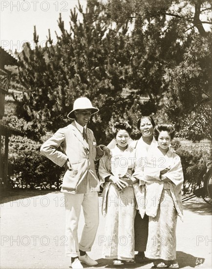 European man with two geisha and their 'okaa-san'. A European man poses outside a Japanese tea house with two female geisha and their 'okaa-san' (the female manager of a geisha house). The girls wear traditional kimonos and their hair is worn up in 'shimada' style. Moji (Kitakyushu), Kyushu Island, Japan, circa 1920. Kitakyushu, Kyushu Island, Japan, Eastern Asia, Asia.