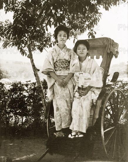 Geisha posing on a rickshaw. Portrait of two geisha from a tea house. They sit side-by-side on a staionary rickshaw, dressed in traditional kimonos and 'geta' (Japanese wooden sandals), with their hair worn up in 'shimada' style. Moji (Kitakyushu), Kyushu Island, Japan, circa 1920. Kitakyushu, Kyushu Island, Japan, Eastern Asia, Asia.
