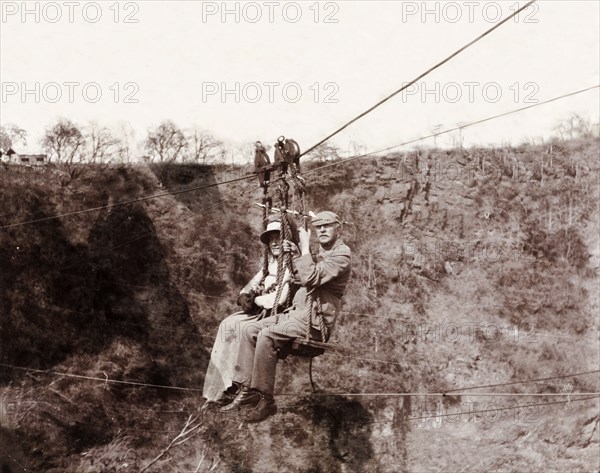 Lady Lawley and Arthur Rhodes cross the Zambezi gorge. Lady Annie Lawley, the wife of Sir Arthur Lawley, Lieutenant-Governor of the Transvaal, and Arthur Rhodes (brother of Cecil John Rhodes) travel across the Zambezi gorge at Victoria Falls on a chair lift attached to a cable system. Matabeleland, Rhodesia (Matabeleland North, Zimbabwe), circa 1904., Matabeleland North, Zimbabwe, Southern Africa, Africa.