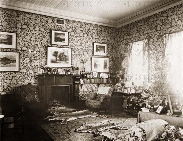 Drawing room of Government House, Bulawayo. The drawing room at Bulawayo's Government House, furnished with a fireplace and bookshelves, and decorated with floral wallpaper, framed pictures and animal skins laid out on the floor. Bulawayo, Rhodesia (Zimbabwe), 1897. Bulawayo, Matabeleland North, Zimbabwe, Southern Africa, Africa.