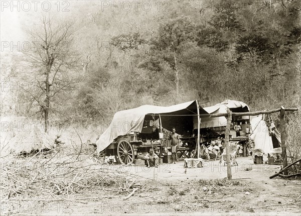 Camp site in the bush. Tents are pitched beside wagons covered with canvas at a camp site in the bush. Two African guides accompany a European man who reclines on a chair lined with animal skin. Near Wankie, Rhodesia (Hwange, Zimbabwe), circa 1898. Hwange, Matabeleland North, Zimbabwe, Southern Africa, Africa.