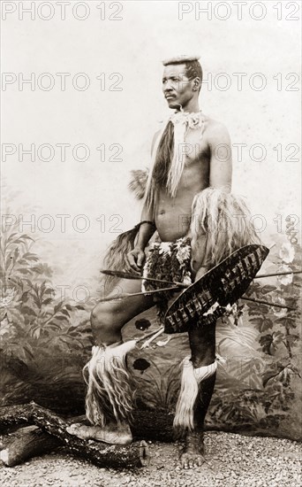 A Zulu warrior. Studio portrait of a Zulu warrior, dressed in traditional Zulu attire and holding a cowhide shield with two 'asagais' or stabbing spears. He stands on gravel in front of a painted backdrop of leafy plants and flowers. South Africa, circa 1897. South Africa, Southern Africa, Africa.